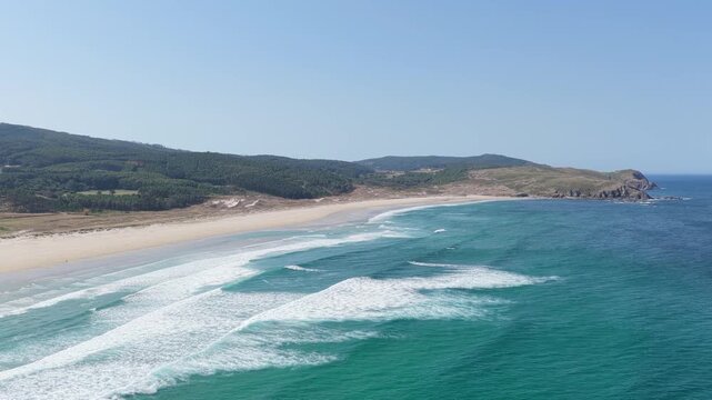 Aerial footage showcasing the sweeping coastline of Praia do Rostro in Spain. The video captures turquoise ocean waves rolling onto a wide, sandy beach bordered by green hills and rugged headlands.
