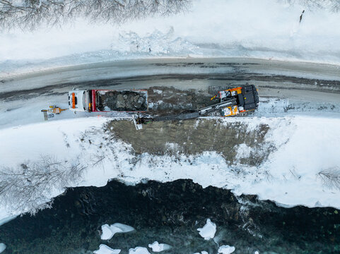 Aerial view of an excavator and a dump truck performing road maintenance on a snow-covered bank beside a river in Valldal, More og Romsdal, Norway.