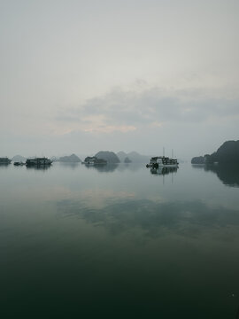 View of traditional cruise boats floating on the calm waters of Ha Long Bay surrounded by limestone karsts under a misty sky in Vietnam.