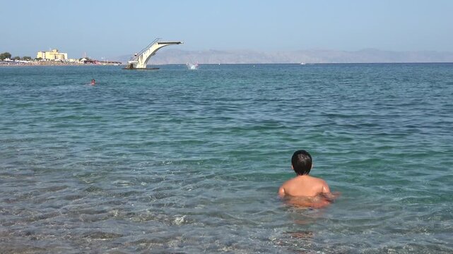 Rhodes Town, Greece - 23 ofOctober 2023: Boy in the sea watching a person dive from the tower at Elli Beach
