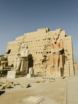 View of the ancient stone statues and the massive pylon wall at the Karnak Temple complex under a clear blue sky in Egypt.