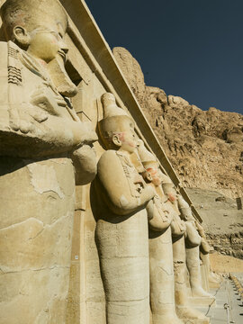 View of the ancient stone statues of Queen Hatshepsut standing in a row at the Mortuary Temple of Hatshepsut against a rocky cliff face under a clear blue sky in Egypt.
