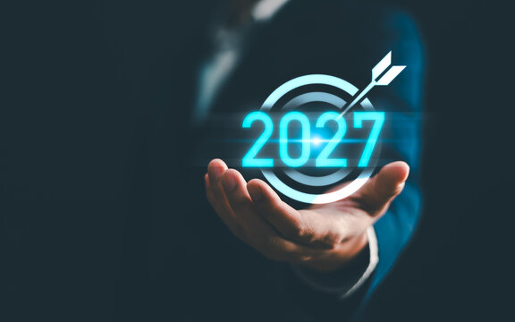 Businessman hand holding glowing year 2027 target icon with arrow on dark background. Concept of business goal achievement, future vision, strategic planning, success and digital technology