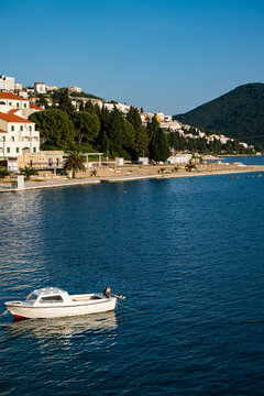 Coastal waterfront of Neum, Bosnia and Herzegovina, with a small white boat on blue Adriatic water, hillside buildings, palms, and mountain