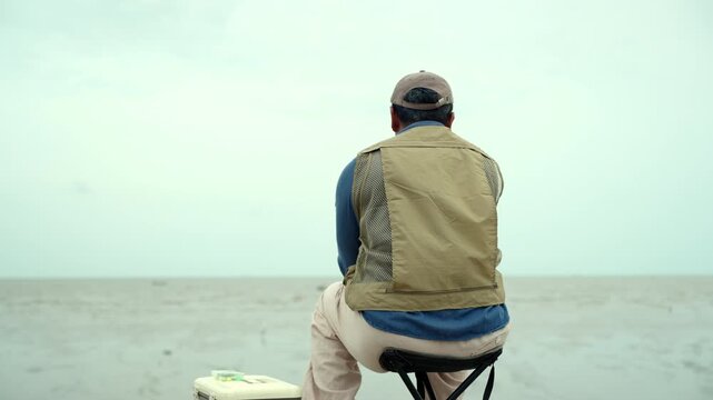 Happy senior man sitting by the sea and fishing with a rod after retirement. Active aging, healthy longevity, outdoor leisure, fulfilling retirement. Life insurance and financial security concept.
