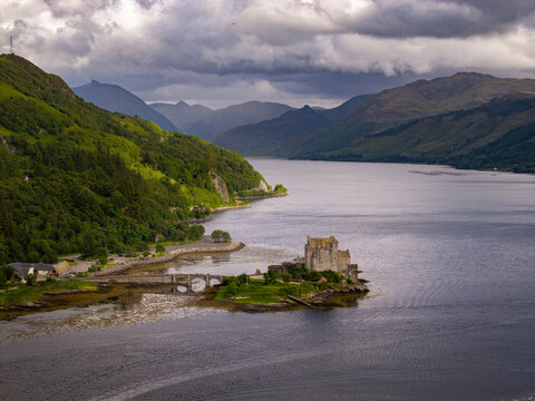 Aerial view of Eilean Donan Castle situated on a small tidal island where three sea lochs meet, surrounded by dramatic mountains and a cloudy sky in Scotland, United Kingdom.