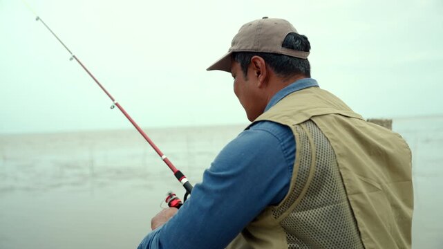 Happy senior man sitting by the sea and fishing with a rod after retirement. Active aging, healthy longevity, outdoor leisure, fulfilling retirement. Life insurance and financial security concept.
