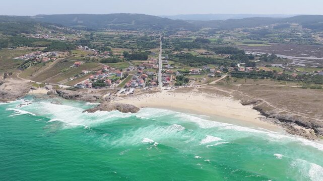 A high-resolution aerial shot showcasing the coastline of Praia de Baldaio in Spain. The scene features turquoise ocean waves crashing onto a wide sandy beach bordered by rocky outcrops.