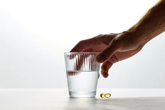 Hand reaching for a glass of water and vitamin capsules on white background