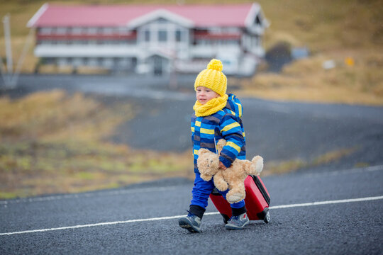 Cute toddler child, boy with teddy bear and suitcase in hand, running on a road in Iceland on a rainy day