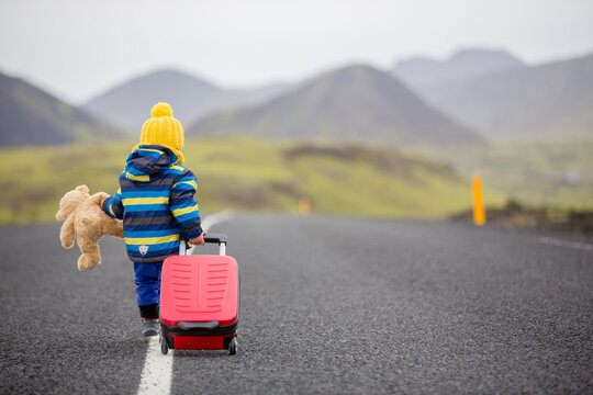 Cute toddler child, boy with teddy bear and suitcase in hand, running on a road in Iceland on a rainy day