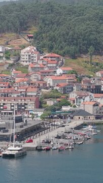 Vertical 9:16 aerial video of the Combarro waterfront in Spain, showing a marina with boats docked along the pier and the hillside village with red-roofed houses in the background.