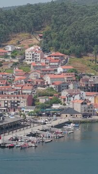 Vertical 9:16 aerial video of Combarro, Spain, showing a picturesque harbor with small boats docked along the waterfront, colorful hillside houses with red-tiled roofs.