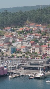 Vertical 9:16 aerial shot of the marina in Combarro, Spain, showing numerous docked sailboats and yachts in the foreground and a hillside village with red-roofed houses.