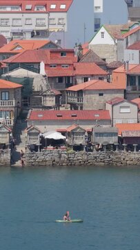 Vertical 9:16 video framing of the picturesque waterfront in Combarro, Spain, showing traditional stone granaries (horreos) lined up along the shore with red-tiled village houses behind.