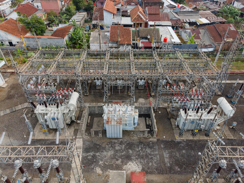 Aerial view of an electrical substation with large transformers and metal power grid structures, near residential houses with tiled roofs in Purwokerto Selatan, Central Java, Indonesia.