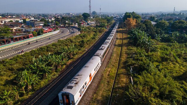 Aerial view of a passenger train traveling along the railway tracks through a lush landscape with a nearby road and urban buildings in Purwokerto, Central Java, Indonesia.