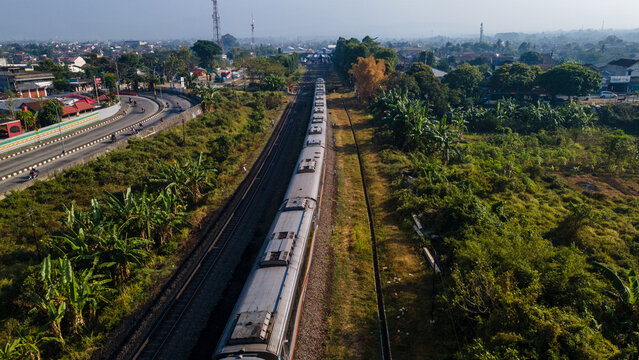 Aerial view of a passenger train traveling along tracks through lush vegetation and trees near a road in Purwokerto, Central Java, Indonesia.