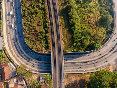Aerial view of a curved road overpass crossing over parallel railway tracks, surrounded by lush green vegetation and scattered buildings in Purwokerto, Central Java, Indonesia.