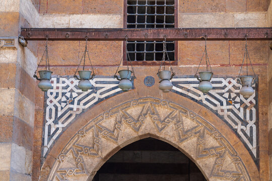 View of the intricate stone entrance and geometric patterns of the Amir Khayrbak Funerary Complex with hanging metal lamps in El-Darb El-Ahmar, Cairo Governorate, Egypt.