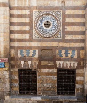 View of the intricate stone facade and circular window of the historic Aqsunqur Mosque featuring traditional ablaq masonry and barred windows in El-Darb El-Ahmar, Cairo Governorate, Egypt.