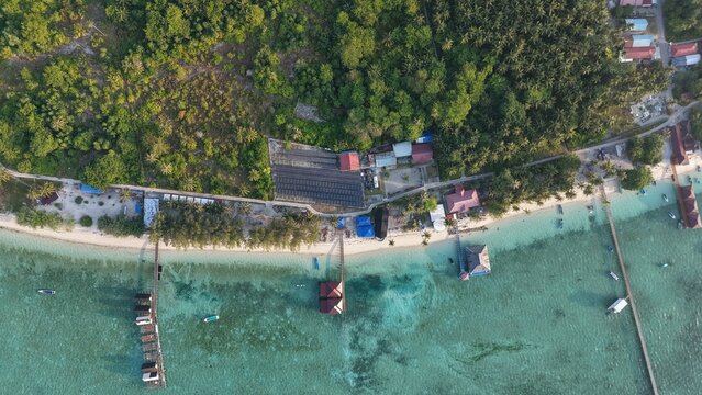 Aerial view of a tropical island coastline in Indonesia featuring wooden piers extending into clear turquoise water, lush green palm trees, and small buildings along the sandy shore.