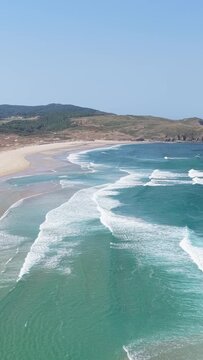 Vertical 9:16 aerial video of Praia do Rostro beach in Spain, showing turquoise waves rolling onto a wide sandy shore with rugged hills in the background.
