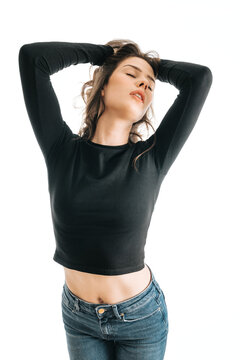 Young woman wearing a black long-sleeve crop top and blue jeans poses with eyes closed. Casual studio portrait shows a relaxed, confident pose with hands behind her head.