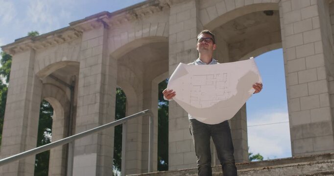 Man wearing glasses walks down the stairs holding a paper plan for the reconstruction of a shopping mall, looking ahead and at the drawing.