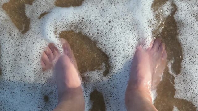 Male feet standing on beach sand as foamy seawater laps over ankles