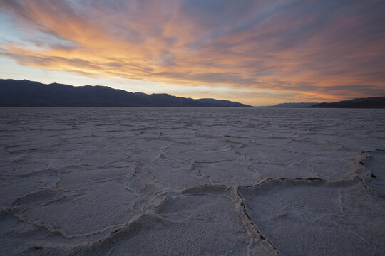 View of the salt flats at Badwater Basin under a dramatic sunset sky with pink and orange clouds over the distant mountains in Death Valley, California, United States.