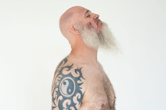 Bald man with a long beard and tattoo on shoulder, side profile.