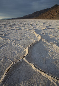View of Badwater Basin salt flats with hexagonal salt crust patterns and rugged mountains in the background under a dramatic sky in Death Valley, California, United States.