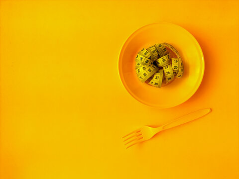 Yellow plastic bowl with measuring tape and plastic fork on yellow background. Represents concepts related to health management, mindful eating, and physical fitness tracking.