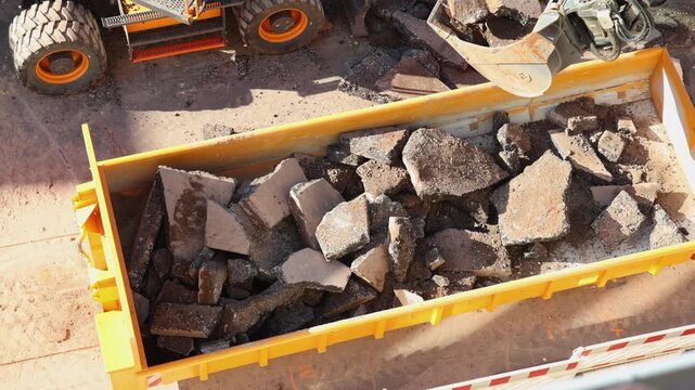 Excavator loads broken asphalt and concrete rubble into construction skip during road demolition. Heavy machinery clears debris from worksite, showing recycling, excavation and infrastructure repair