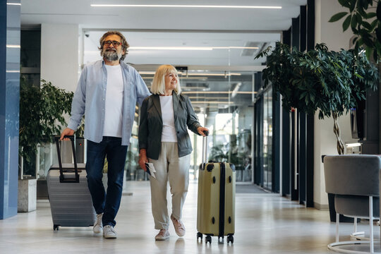 Mature couple walking together in a modern hotel lobby, carrying luggage, with large windows and indoor plants visible in the contemporary setting