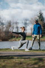 Obraz premium A girl performs a deep lunge while another girl acting as a trainer watches and guides. The outdoor workout takes place on grass near a lake in a park setting.