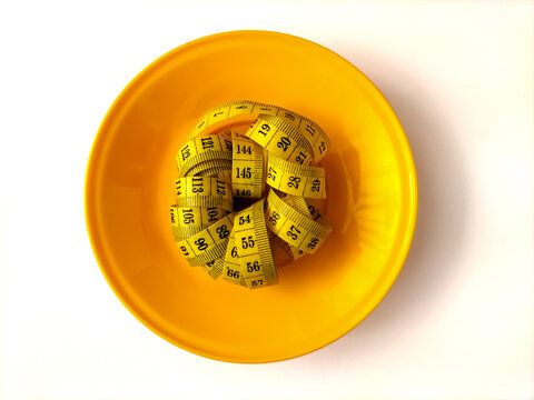 Coiled yellow measuring tape served inside a yellow plastic plate. This minimalistic composition visually highlights strict dieting, obesity prevention, healthy portion limits, and fitness goals