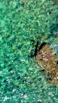 Vertical loopable top down view of a rock surrounded by crystal clear turquoise sea with gentle waves creating a seamless tropical ocean background.