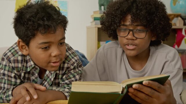 African American teenager and kid lying down and reading book together, turning pages and discussing story. Ideal for educational marketing, literacy campaigns, and family storytelling