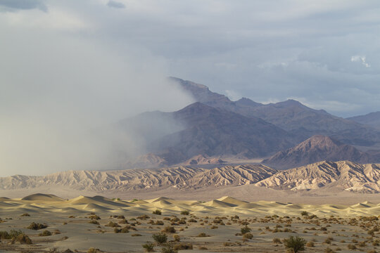View of Mesquite Flat Sand Dunes with wind-blown sand, rugged mountains in the background, and dramatic cloudy sky in Death Valley, California, United States.