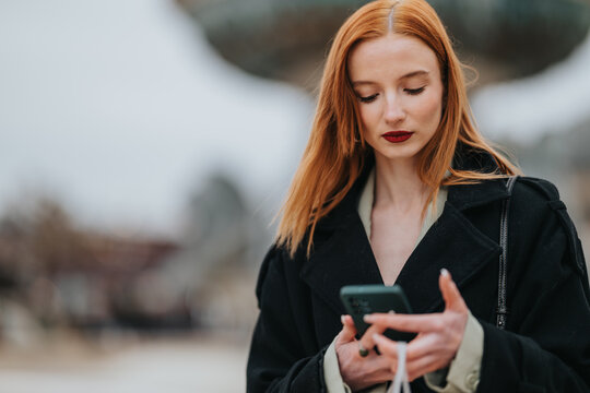 Young red-haired girl looks at her smartphone while standing outdoors in a coat. She checks her phone with a thoughtful expression against a blurred urban background.