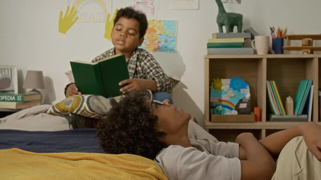 African American kids studying in bedroom, one boy reading book aloud while other boy lying down listening and dreaming. Ideal for educational campaigns, childhood development, or family promos