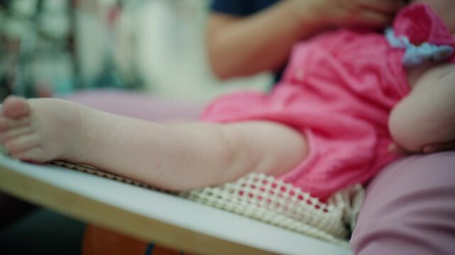 Baby sitting on a wooden table, wearing a pink dress, with adult hands providing gentle support. Childhood and infancy care concept