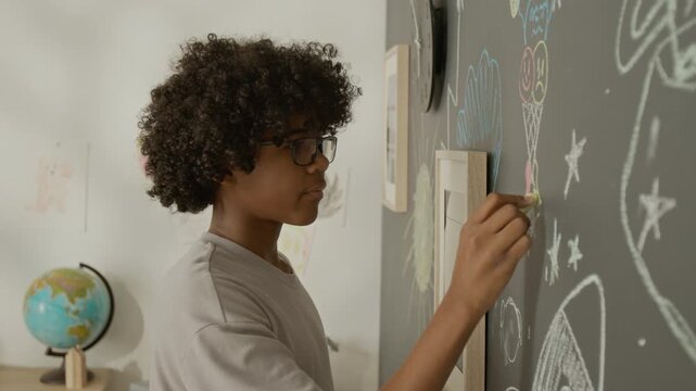 African American teenager wearing glasses drawing on blackboard with colored chalk. Footage is perfect for educational campaigns, creative learning promotions, or youth development advertisements