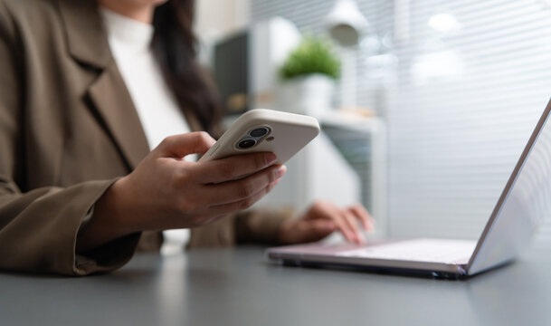 Businesswoman using a smartphone while working on a laptop, enabling mobile communication, digital workflow, and online collaboration in a modern professional workspace.