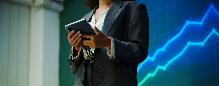 Businesswoman holding a tablet during a financial presentation with rising market graphs on a large screen, representing stock market analysis, investment growth, trading, strategy, and finance.