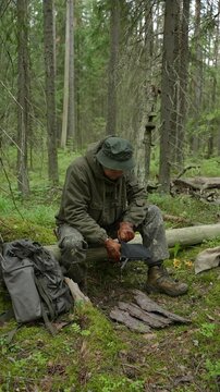 Man in tactical clothing sitting on a log and unzipping a pouch to take out a portable pocket twig stove. Concept of bushcraft camp cooking, wilderness survival gear, wild camping equipment and outdoo