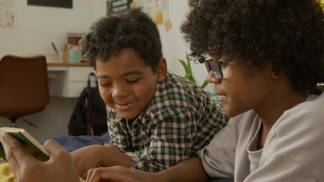 African American teenager and child lying on bed, reading book together and laughing. Ideal for educational campaigns, family bonding promotions, and literacy advertisements