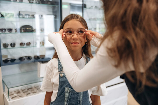 Young girl trying on glasses with assistance from an adult in an eyewear store, display of sunglasses and optical frames visible in the background